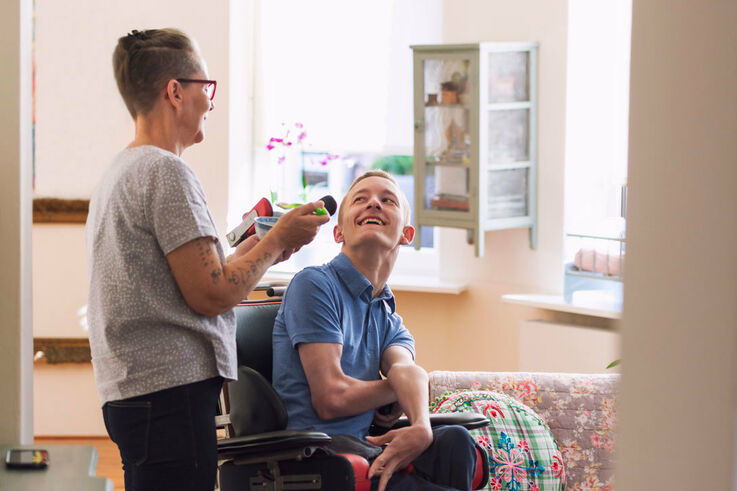 Eine Diplom-Sozialbetreuerin aus dem Bereich Behindertenarbeit steht neben einem jüngeren Mann im Rollstuhl und beide lächeln sich an (Bild: FSW)