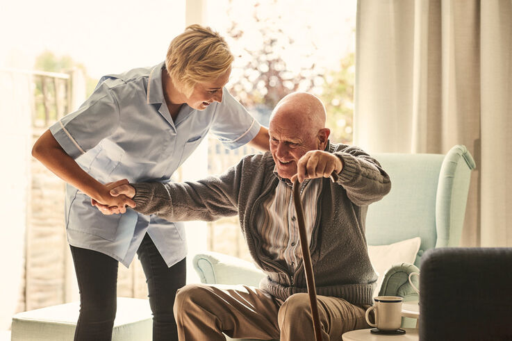Eine Heimhelferin hilft einem älteren Mann mit einem Stock in seiner Hand beim Aufstehen von einem Sessel (Bild: FSW)